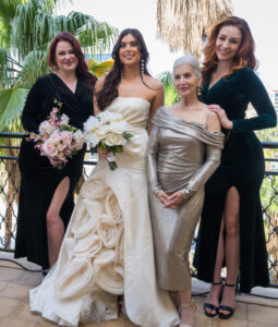 A balcony group portrait by High-Tech Studios in Los Angeles, featuring a bride in a cream gown, her mother in a metallic gold dress, and two bridesmaids in dark velvet gowns backed by palm trees.