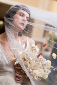 Close-up bridal portrait by High-Tech Studios in Glendale, CA, showing a bride looking up with a delicate white veil flowing across the frame while holding a textured neutral floral bouquet.