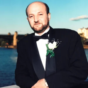 A formal portrait by High-Tech Studios in Los Angeles, showing a groom in a classic black tuxedo with a white rose boutonniere standing by the water with a bridge in the background.