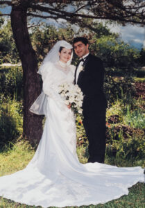 An outdoor wedding portrait by High-Tech Studios in Pasadena, featuring a newlywed couple standing in a field in front of large redwood trees in classic wedding attire.