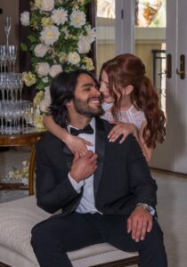 A candid photograph of a modern couple in a formal embrace. A man in a black tuxedo is seated, while a woman in a white lace dress hugs him from behind, her head resting on his shoulder. An elaborate white flower arrangement and a champagne glass tower are visible in the background.