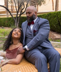A seated photograph of an African American couple in an outdoor garden with palm trees. A man with a beard, wearing a grey suit and a red velvet bow tie, is seated on a light woven bench. A woman, looking up at him with a smiling gaze, is seated below, wearing a rust-colored velvet dress.