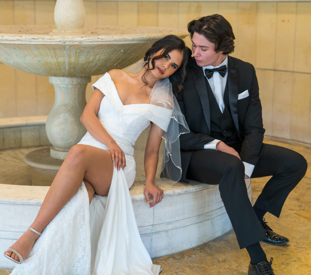 A high-resolution wedding portrait of a bride and groom sitting on the edge of a tiered stone fountain in a Burbank hotel lobby. The bride leans her head on the groom’s shoulder. Captured by High-Tech Studios.